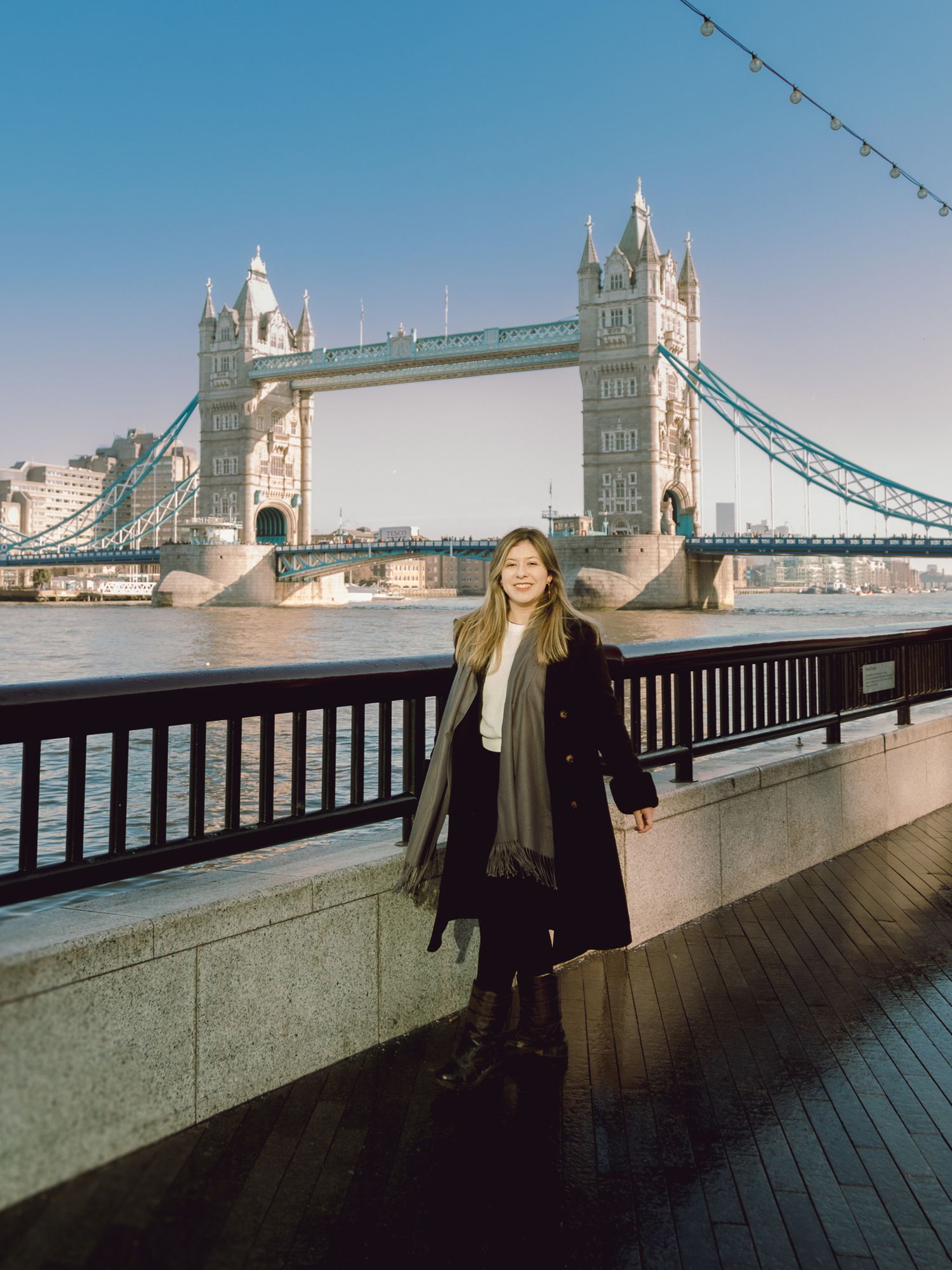 Person in winter coat poses against London's Tower Bridge on a clear blue day.