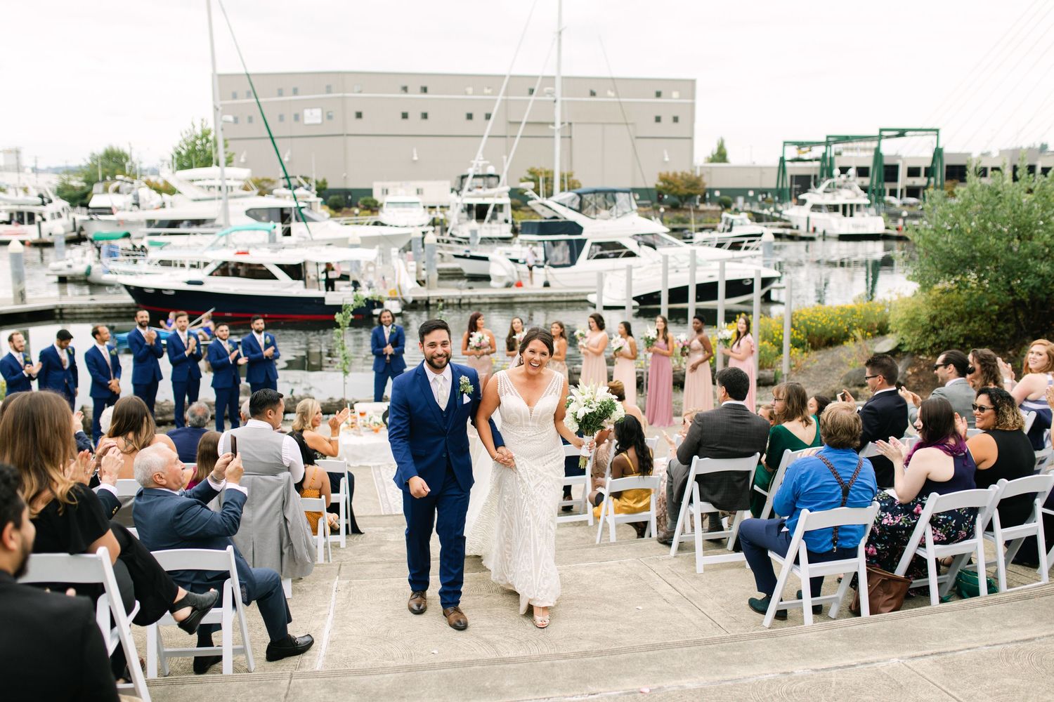 Outdoor waterfront wedding ceremony at a marina with boats and guests seated in white chairs by the dock.