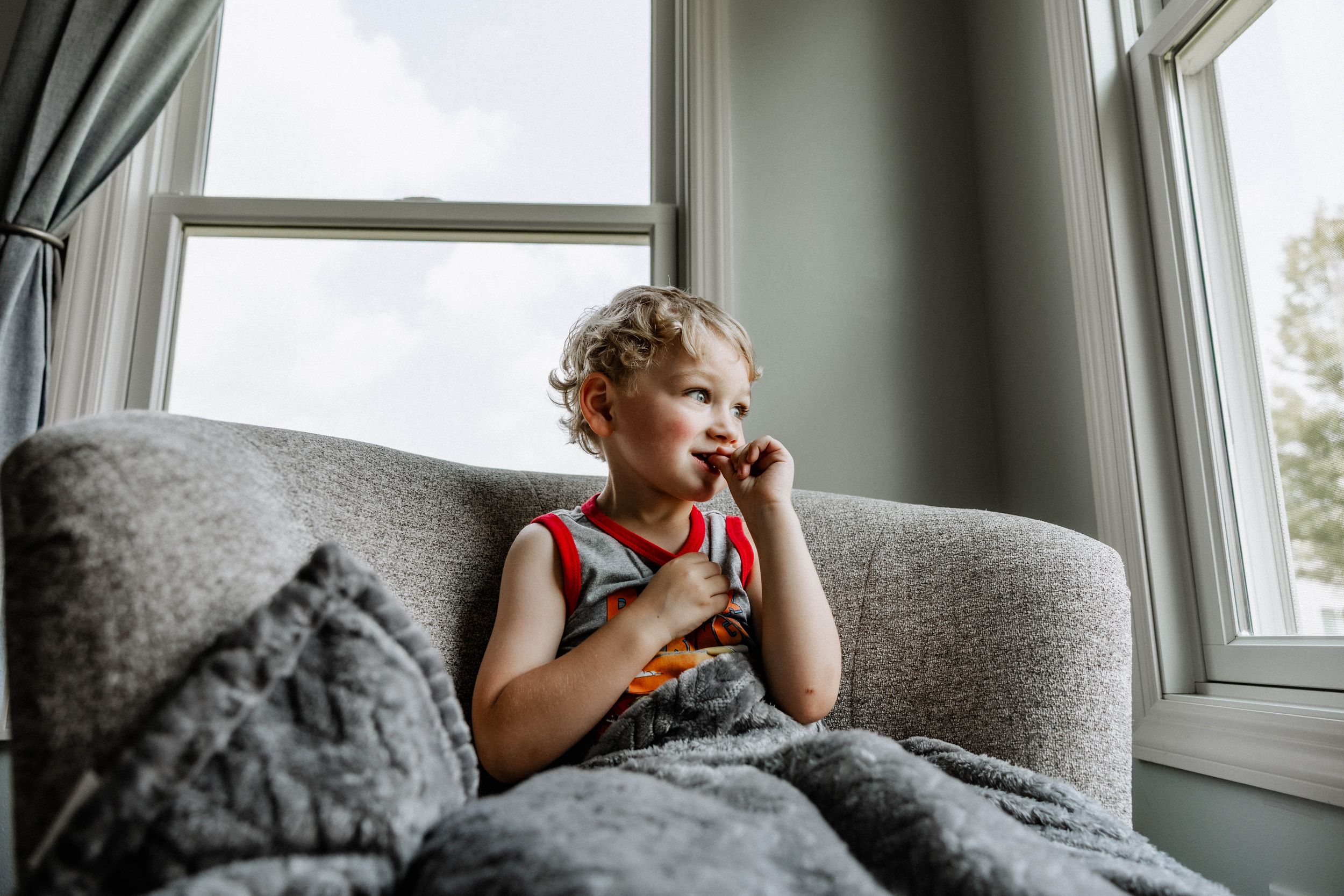 Young child in sleeveless top sits on gray couch by window under blanket while holding electronic device.