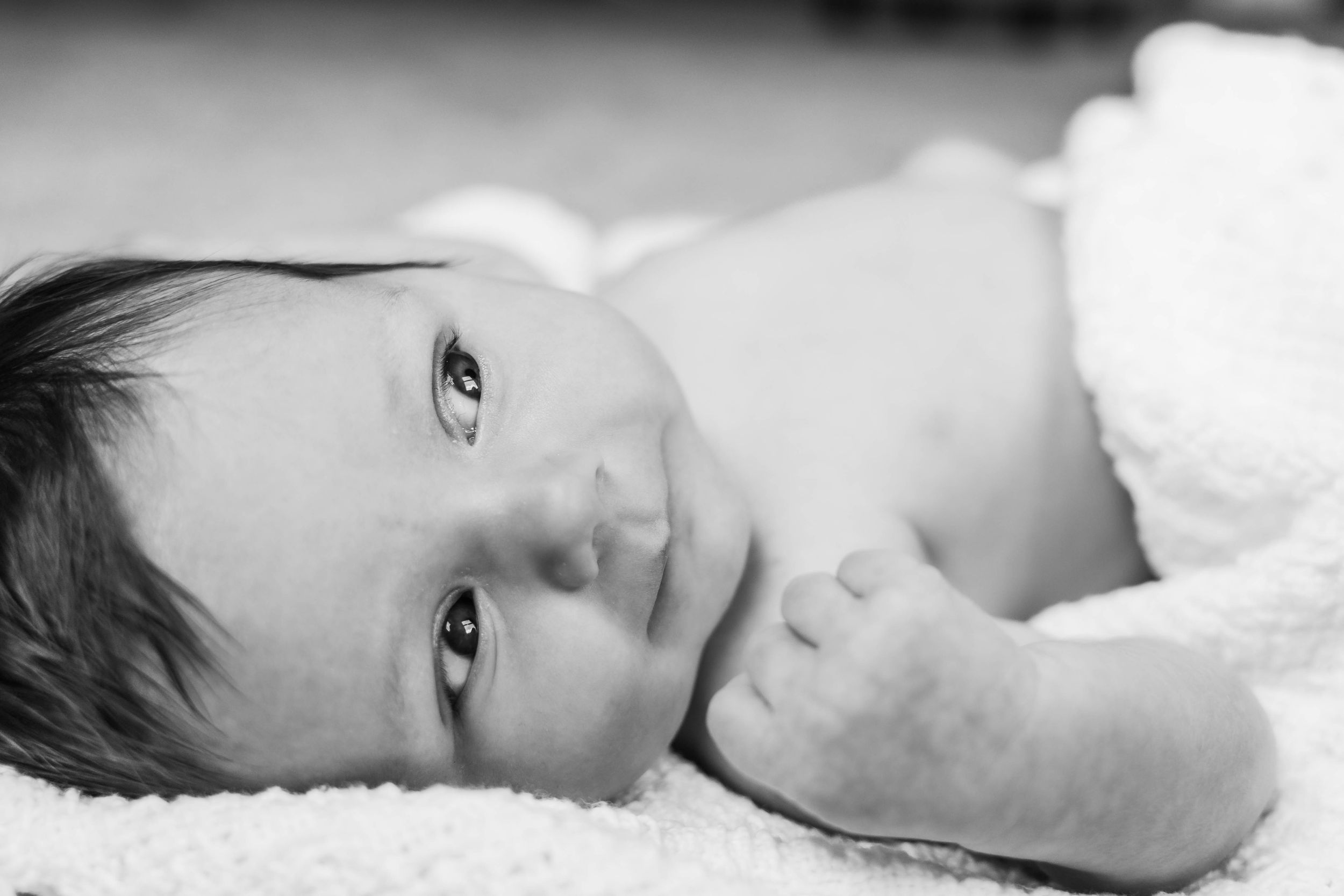 A small baby lies on white bedding in a black and white portrait capturing their serene expression.