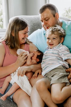 A loving family shares joyful laughter while sitting together on a cozy gray couch near a bright window.