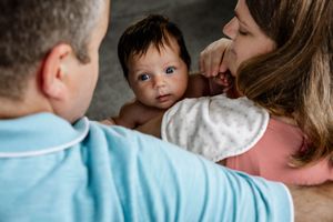 A baby gazes intently while being held by two adults wearing blue and pink shirts in an intimate family moment.