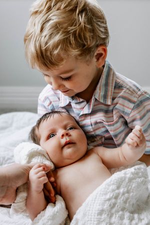 A young child leans over a baby lying on white blankets, sharing a tender moment together in soft lighting.