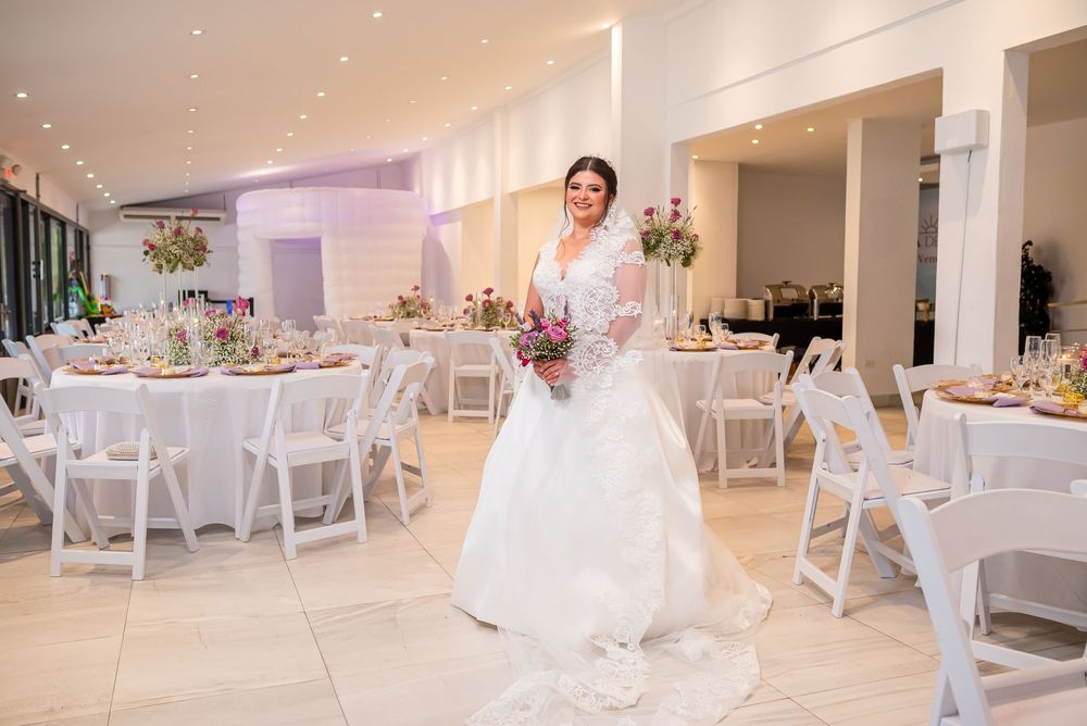 A person in an elegant white wedding gown stands in a beautifully decorated reception venue with round tables and chairs.