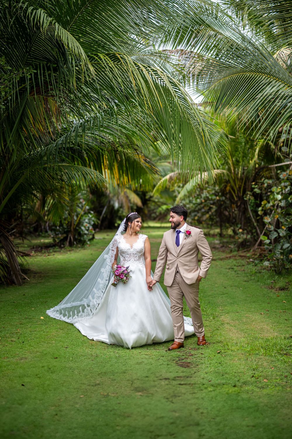 A romantic wedding photoshoot in a lush tropical garden with palm trees as newlyweds walk along a grassy path.