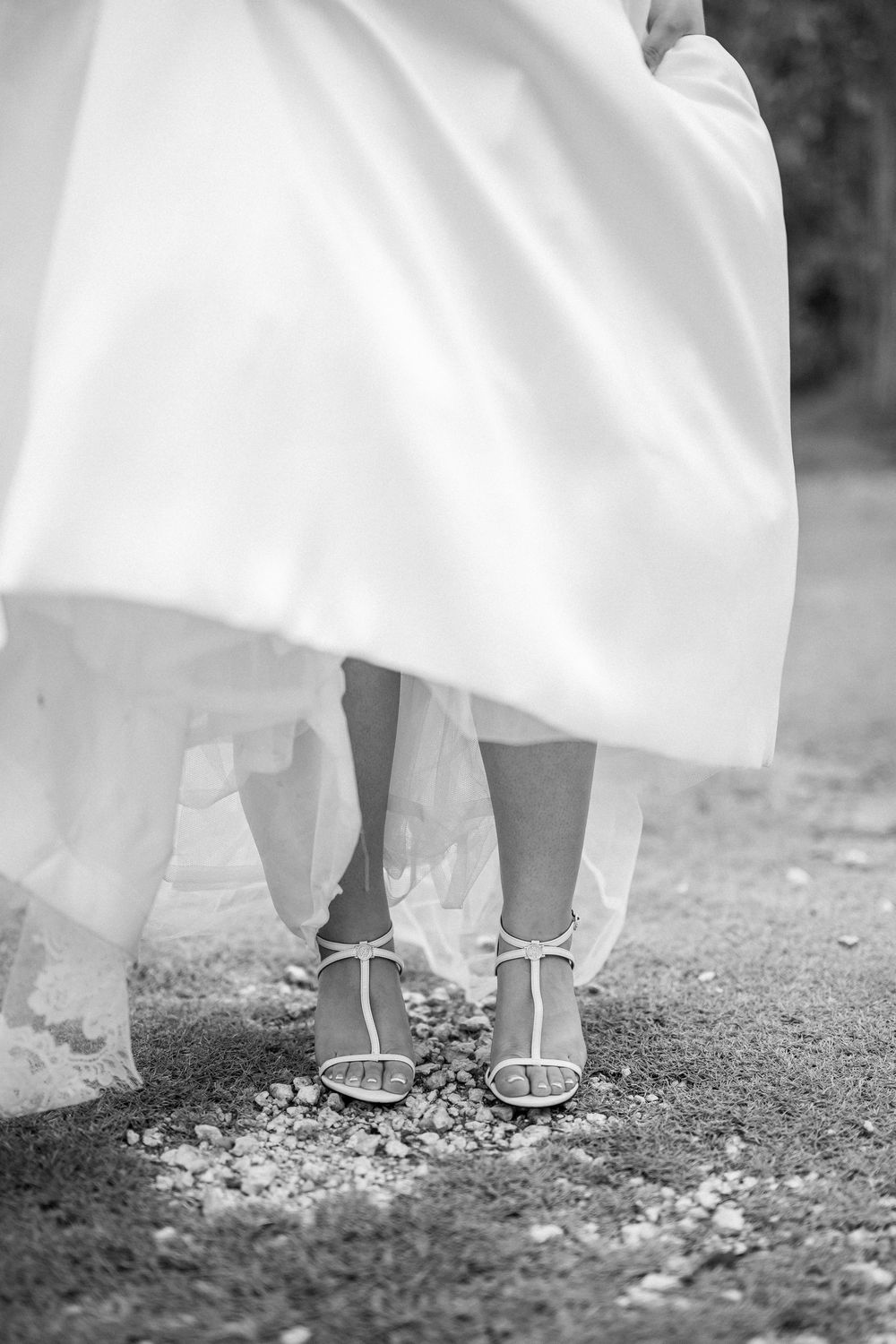 Close-up black and white photo of elegant bridal shoes and hem of white wedding dress on gravel ground.