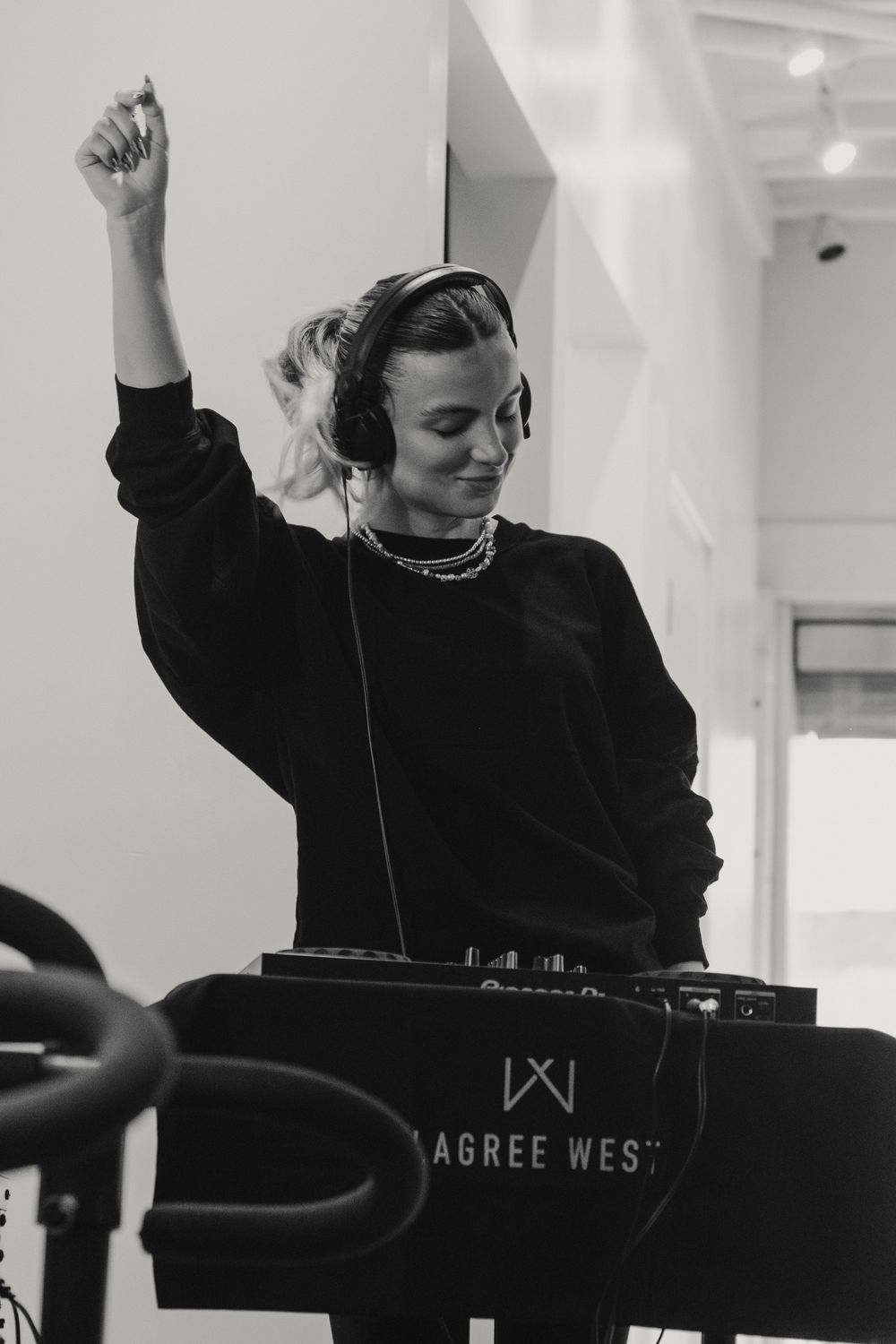 DJ in a black sweater raises arm while performing behind turntables at an event in black and white photography.