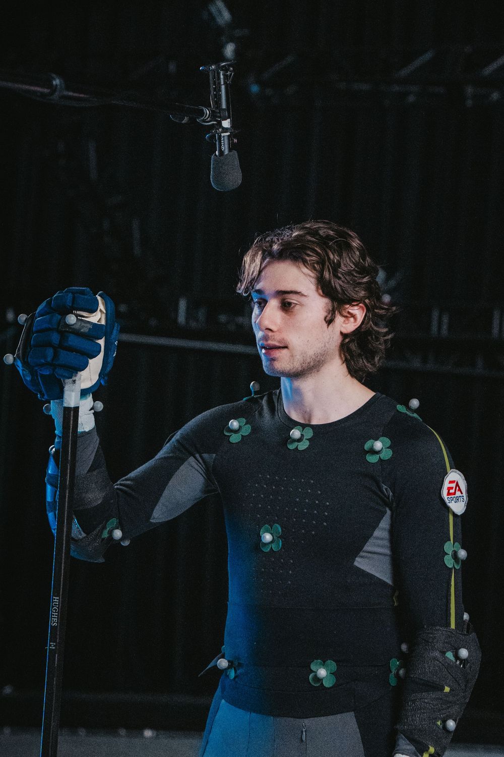 Motion capture performer in black suit with tracking markers holds motion capture equipment in studio setting.