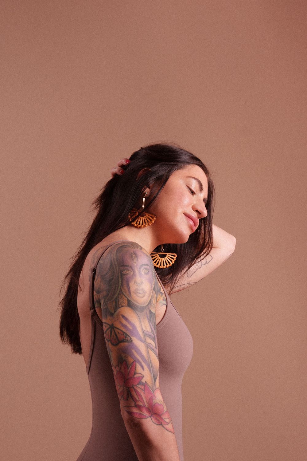 Side profile of a model with ornate sleeve tattoos and golden fan earrings posing against a mauve background.