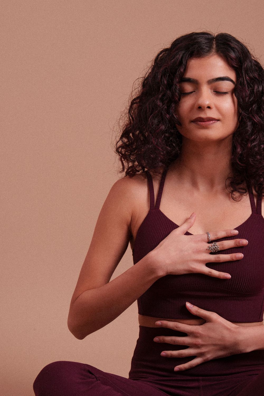 Person with curly dark hair in burgundy activewear practicing mindful meditation against warm brown background.