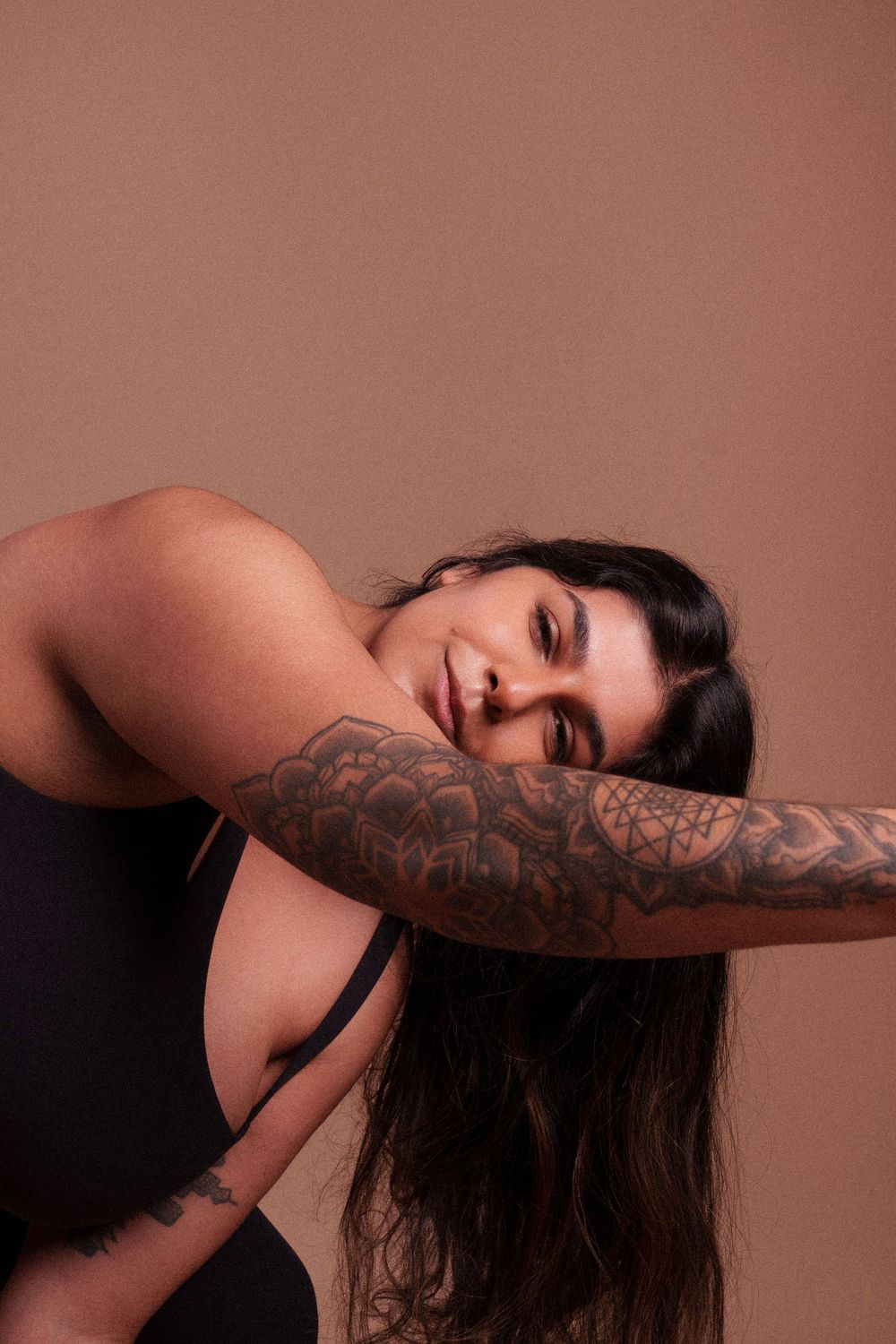 A person with long dark hair and detailed floral tattoo sleeve poses gracefully against brown backdrop.