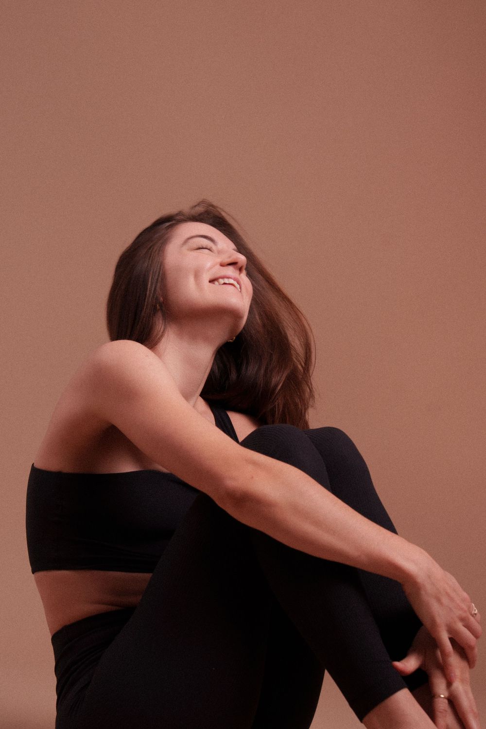 Person in black athletic wear laughing joyfully while sitting against brown background, demonstrating carefree emotion.