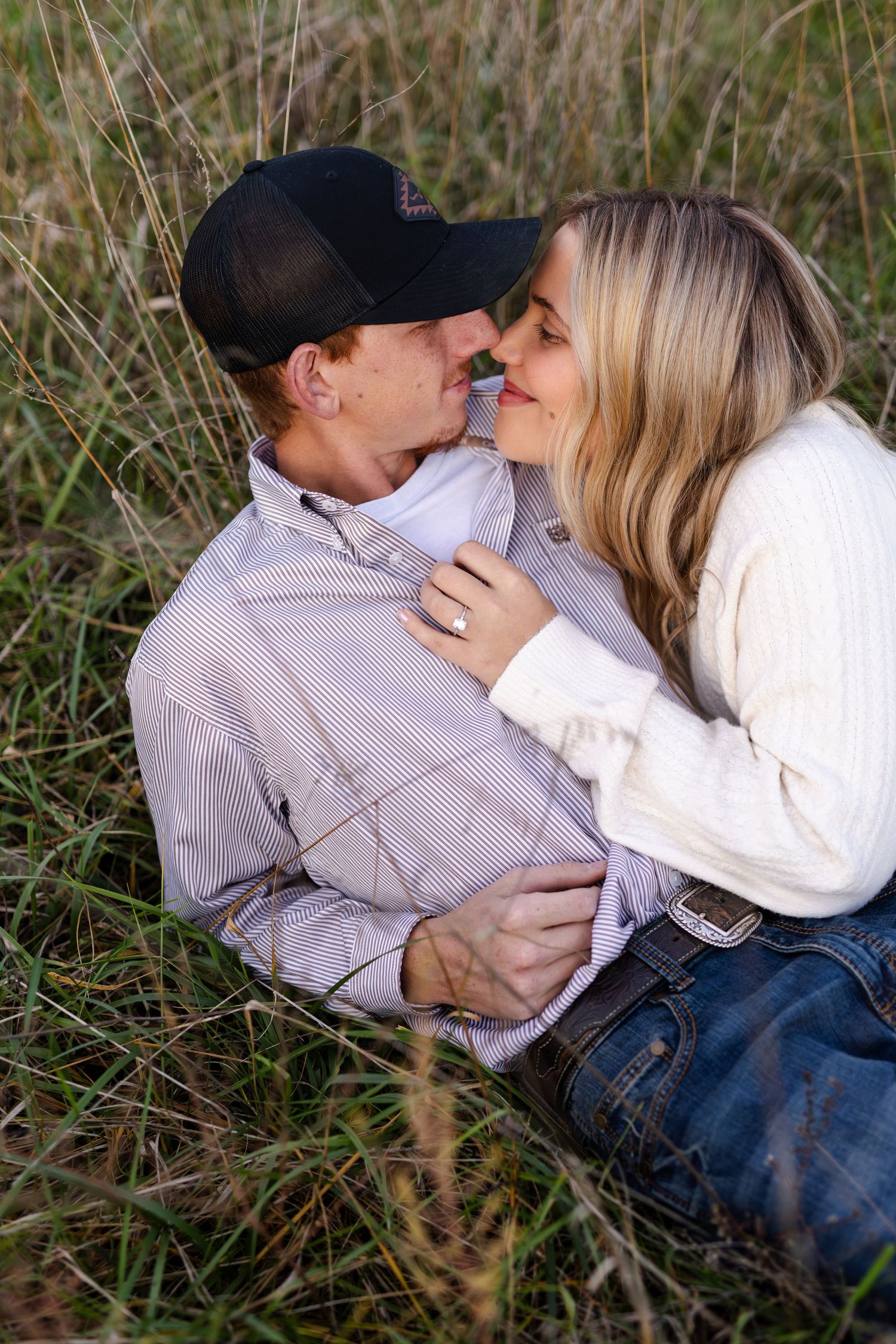 Romantic couple sitting in tall grass sharing a kiss during a warm outdoor engagement photo session.