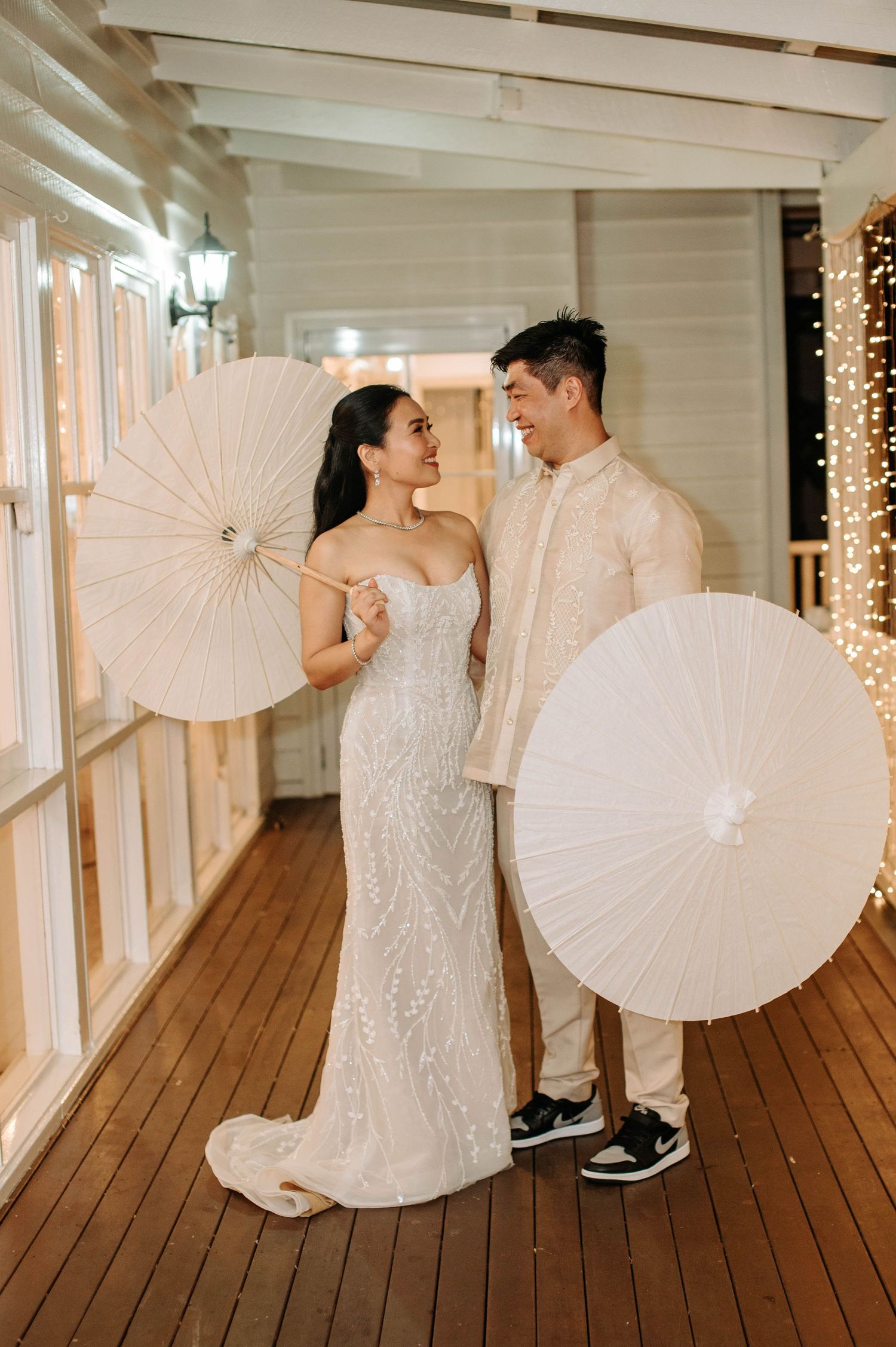 Couple posing with white parasols in a beautifully lit indoor venue with wooden floors and string lights.