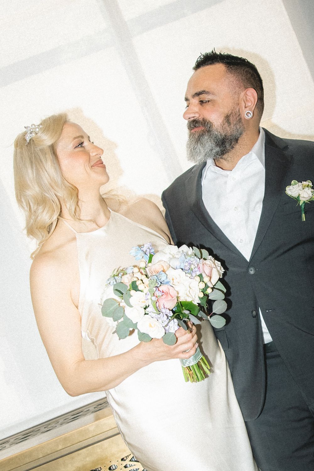 Bride and groom share a tender moment, she holds a pastel bouquet, he wears a dark suit with boutonniere.