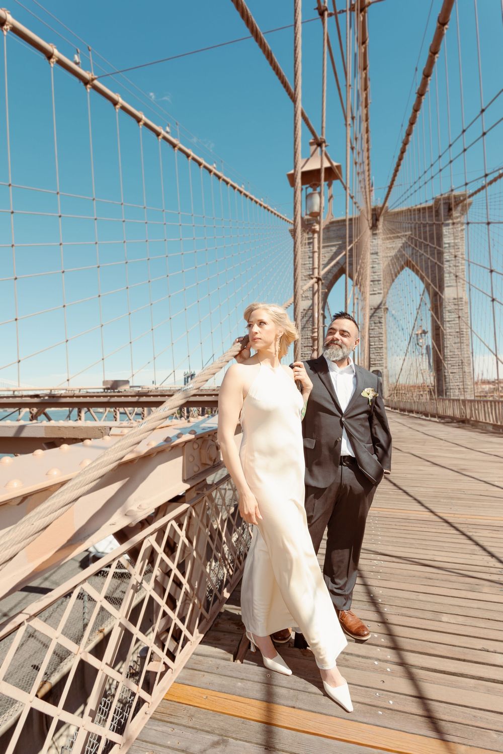 Bride and groom posing on Brooklyn Bridge on their wedding day, with blue sky and bridge cables behind them.
