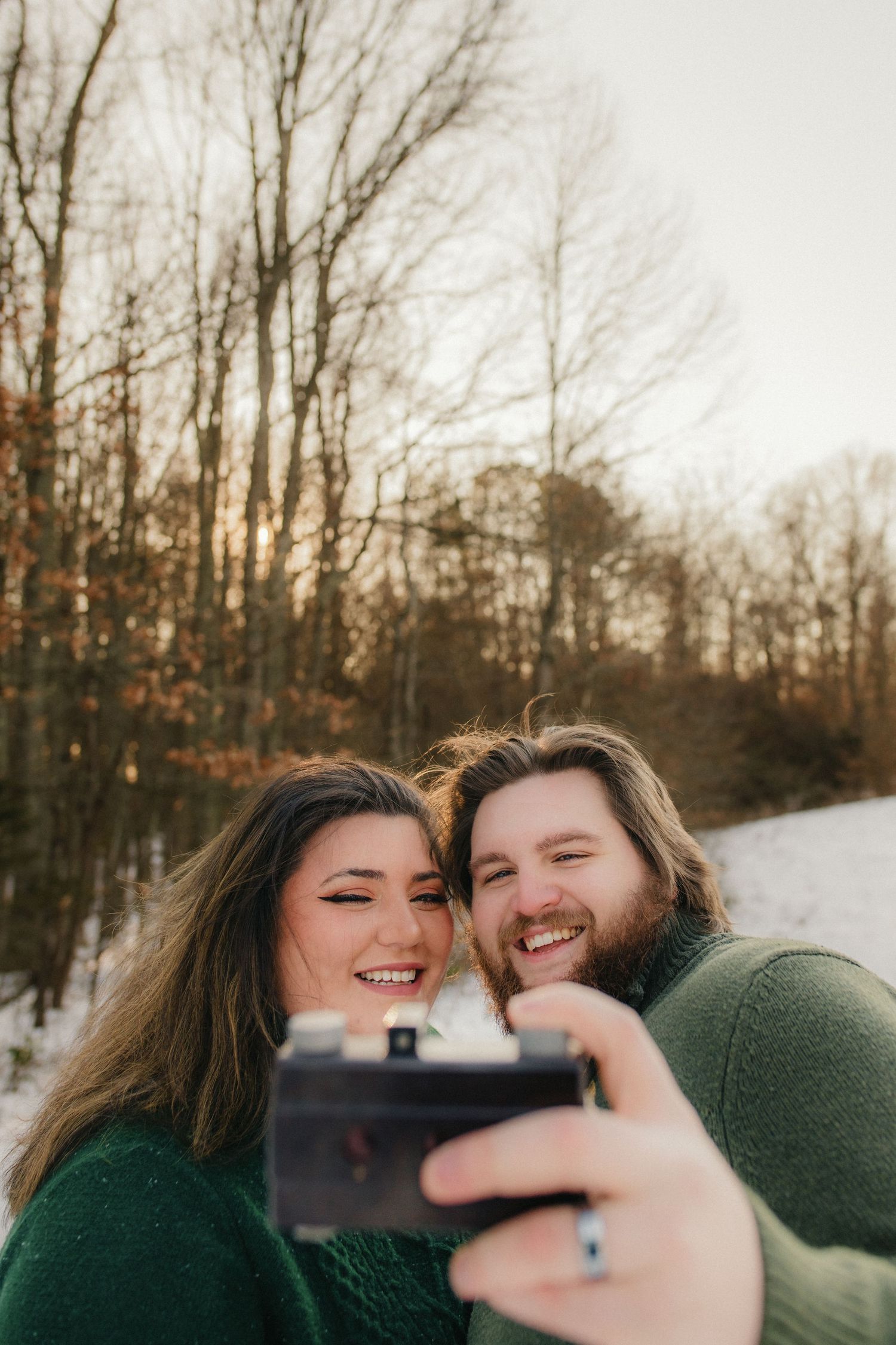 A couple takes a selfie together in the woods during golden hour wearing matching green sweaters.