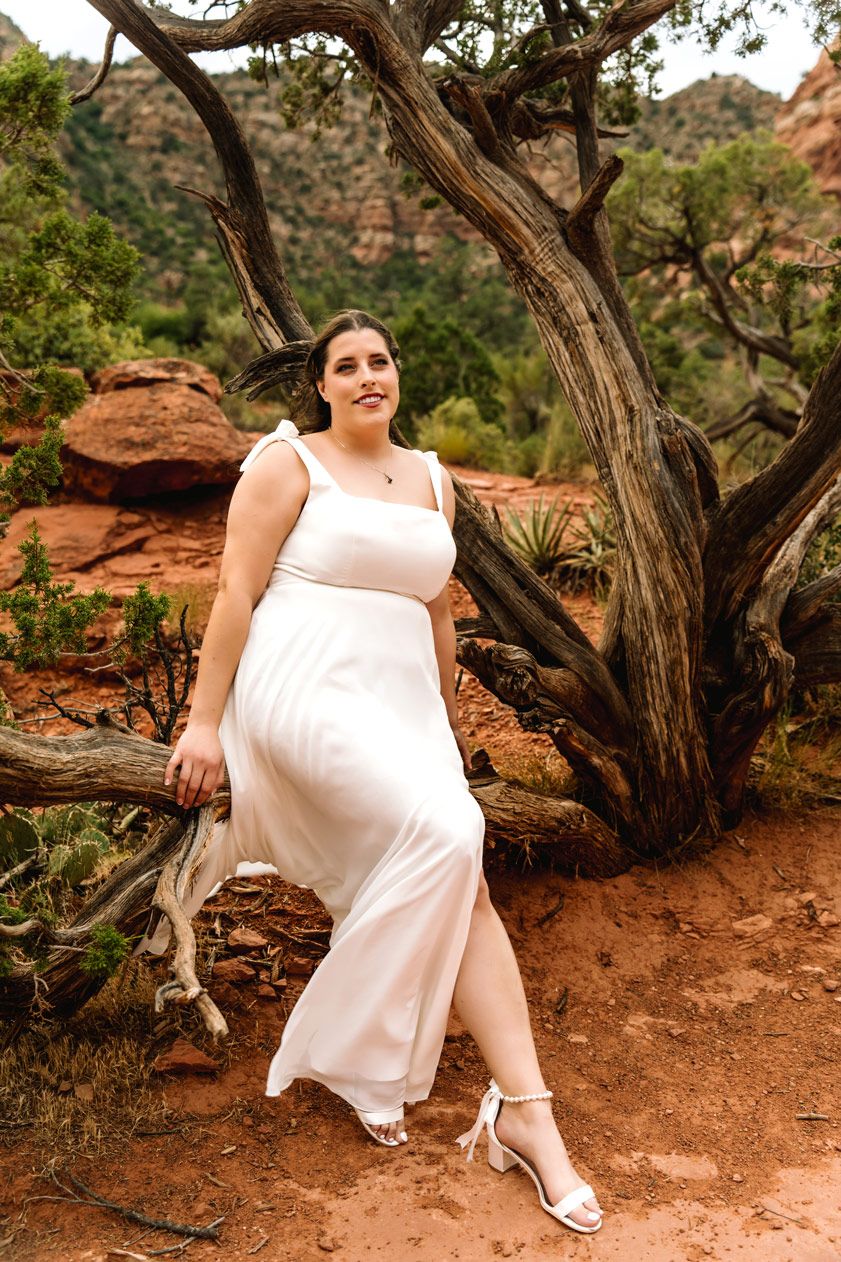 Bride poses elegantly by twisted juniper tree on Sedona red rock trail.