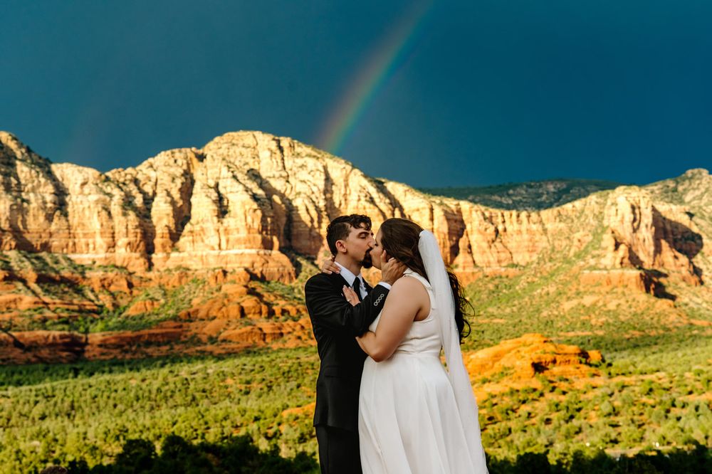 Wedding couple poses beneath a vibrant rainbow against dramatic Sedona red rock cliffs.