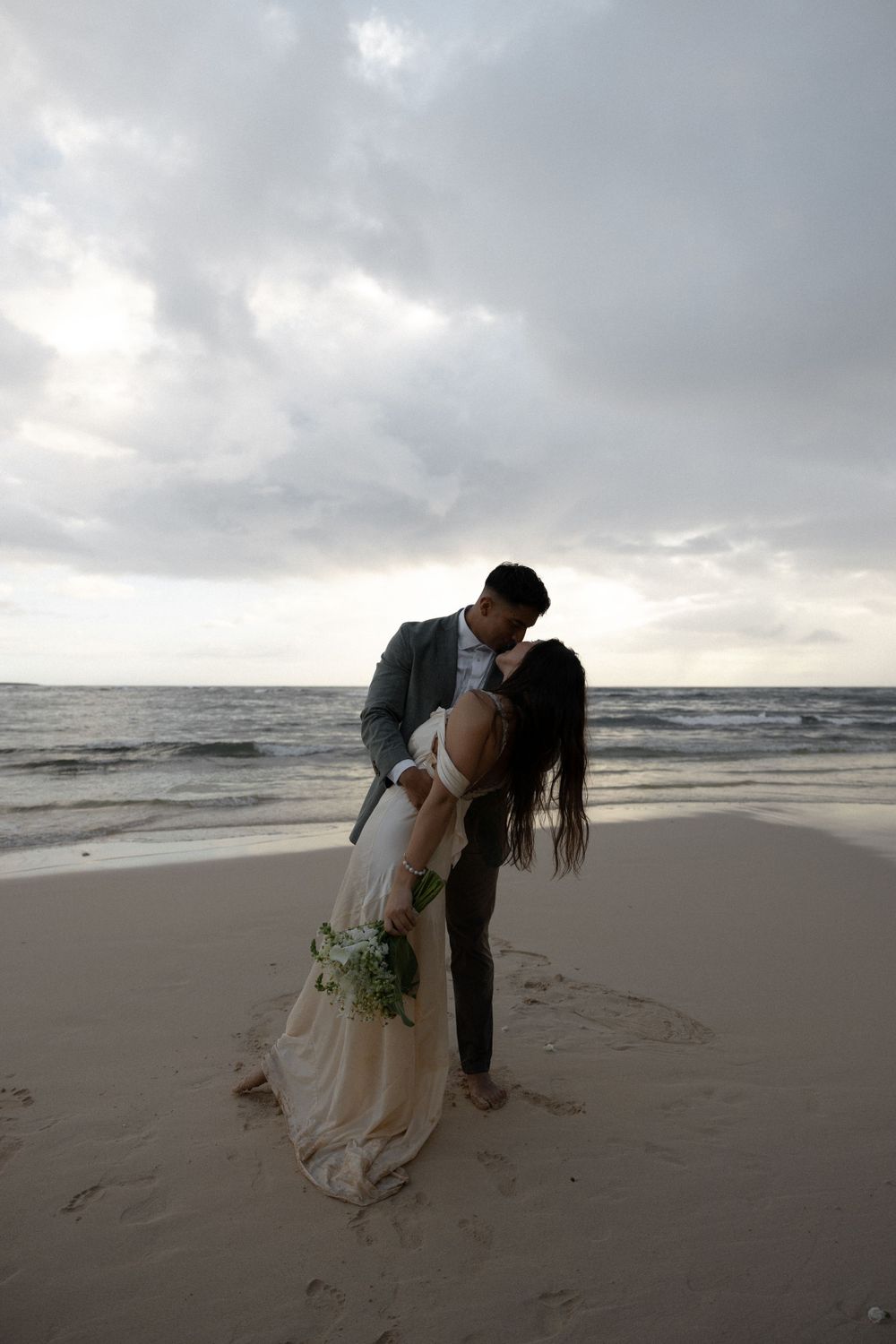 A romantic dip pose on the beach with dramatic cloudy skies and ocean waves in the background.