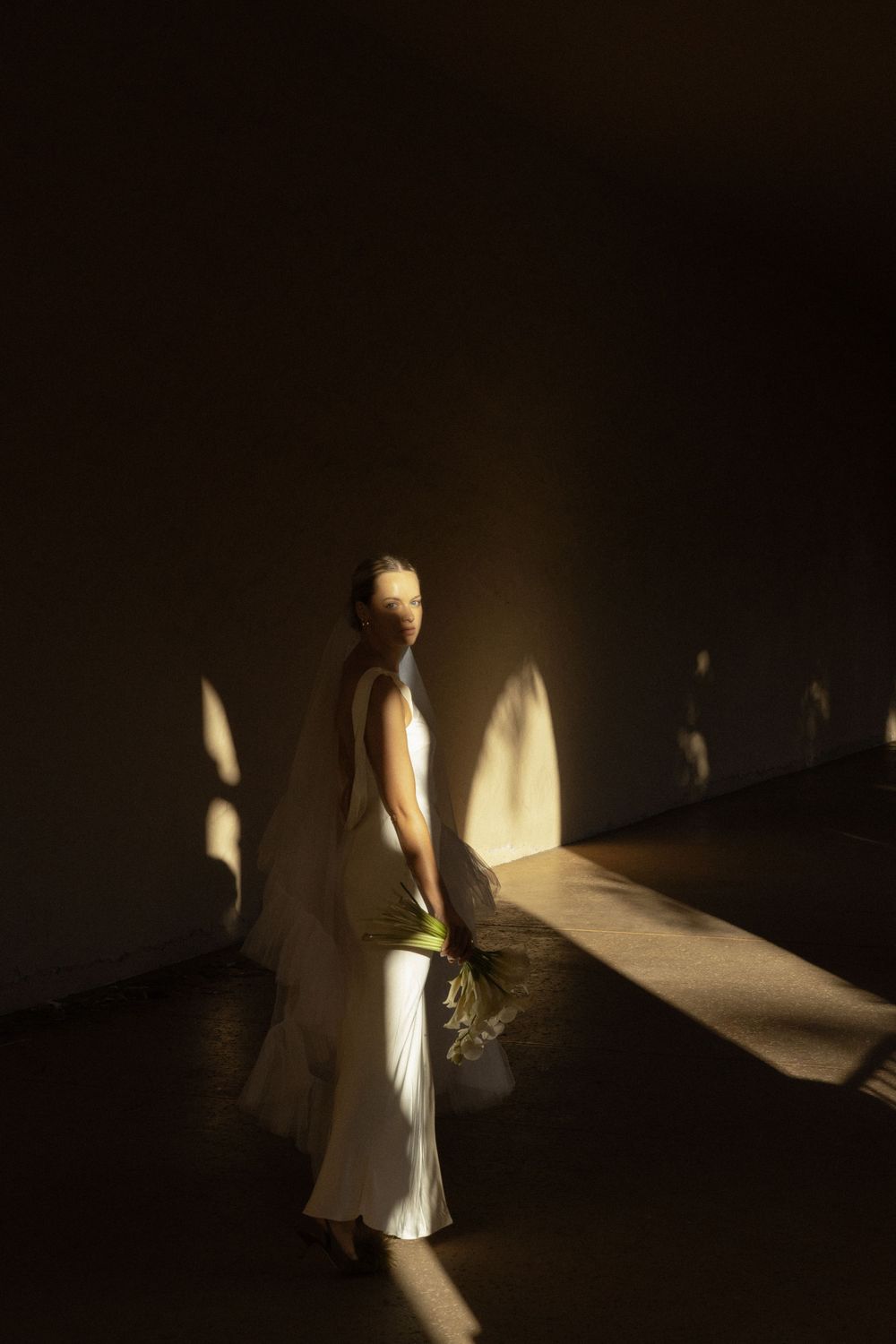 A bride in a flowing white dress standing in dramatic shadows and light.