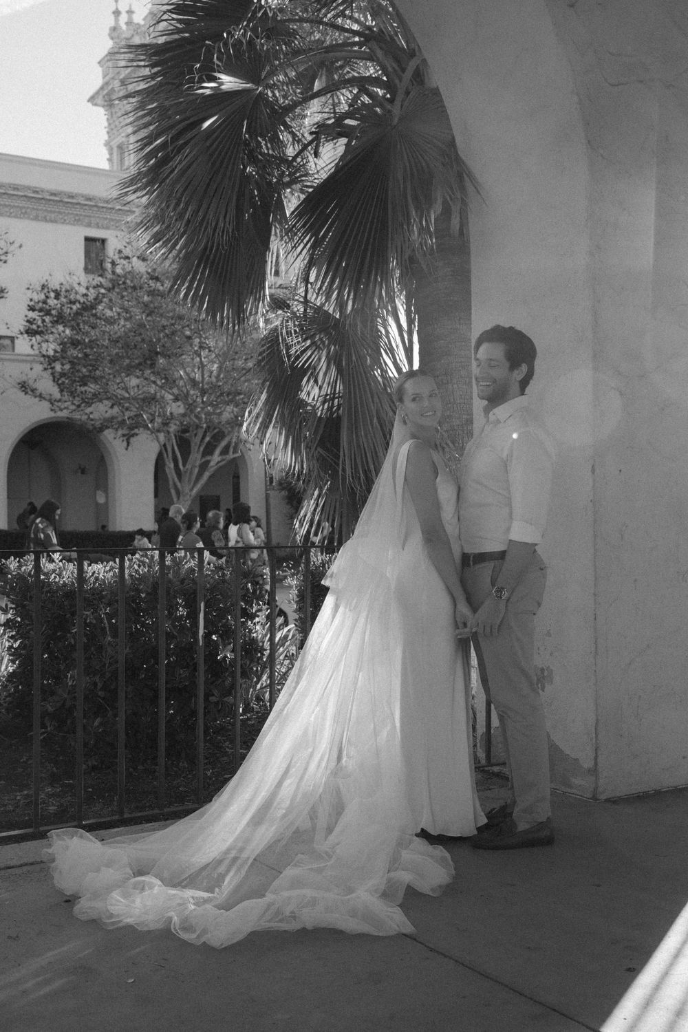 A black and white photo of a couple by a palm tree in classic wedding attire.