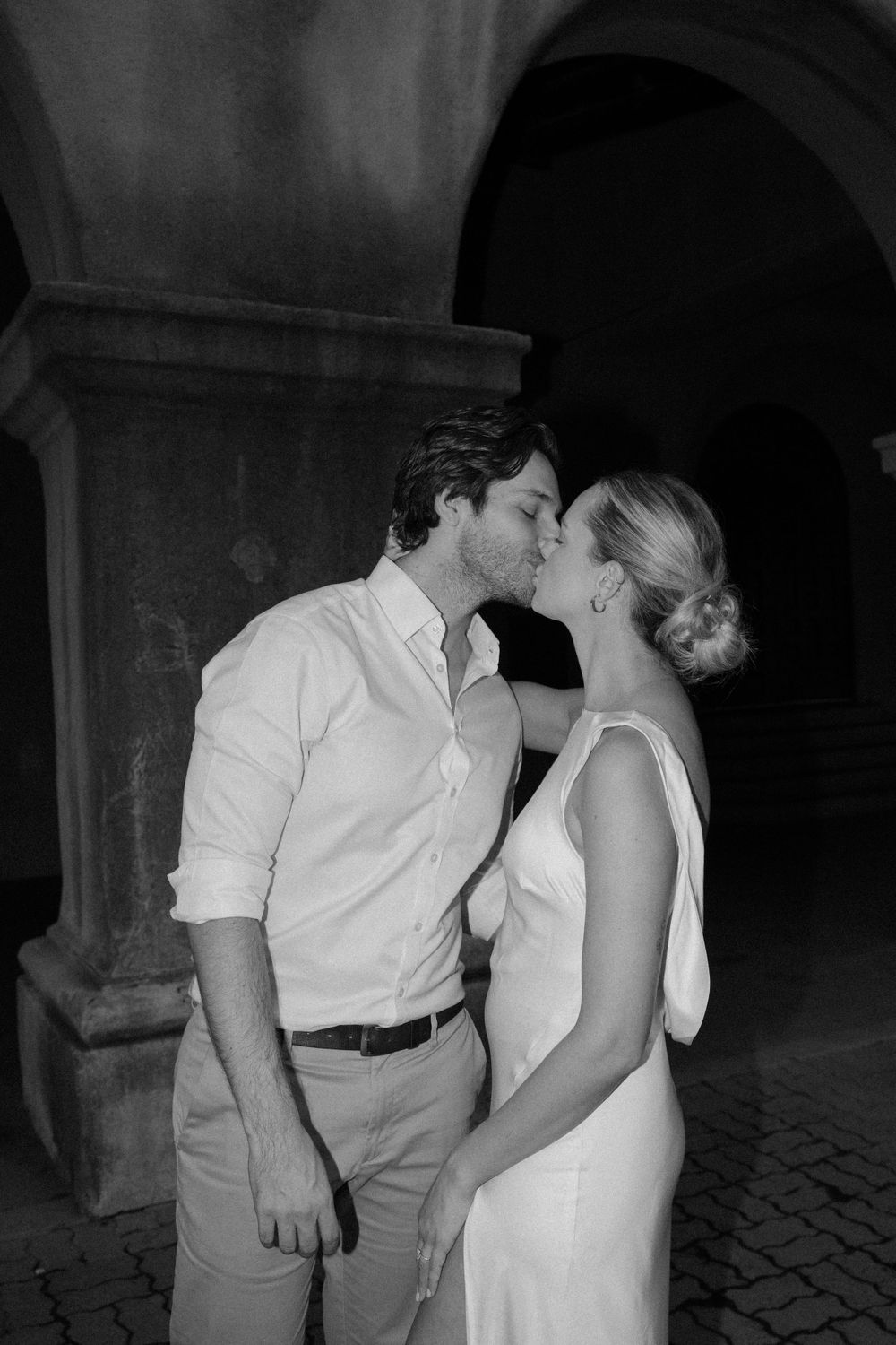 A romantic moment captured between a couple in formal wear under an archway, black and white flash photography.