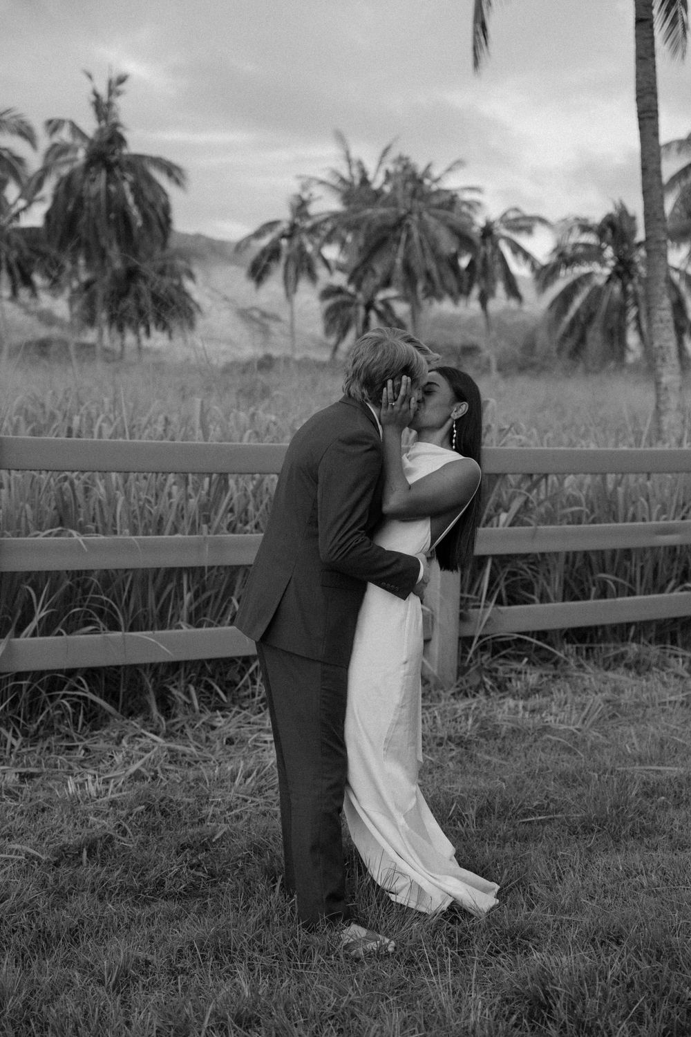 A black and white photo of a couple embracing by a fence with palm trees in the background.