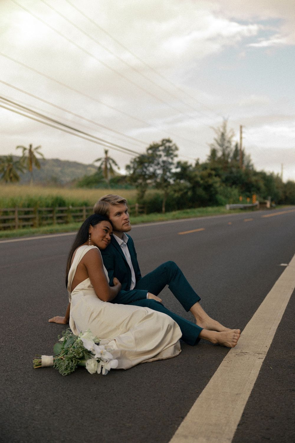 A couple sits together on a rural road with mountains and palm trees in the background.