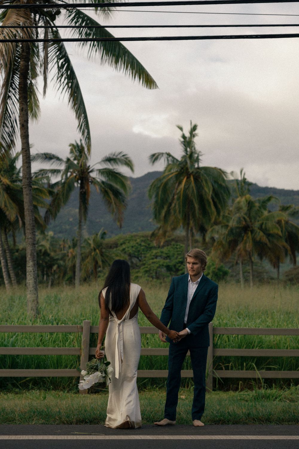 A couple stands together on a road with tropical mountains and palm trees in the background.