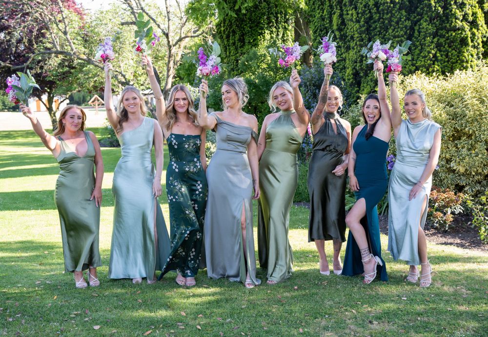 Bridesmaids in sage and blue dresses pose together on sunny garden lawn.