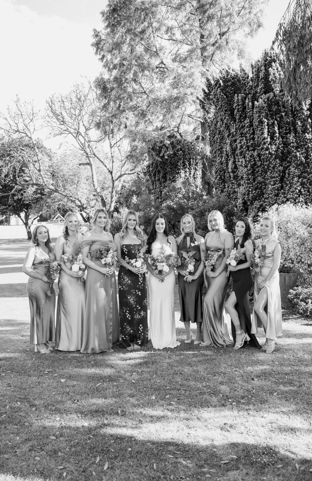 Black and white portrait of bridal party standing together outdoors under trees.