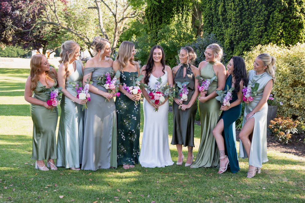 Wedding party in sage green dresses poses for group photo in garden setting.