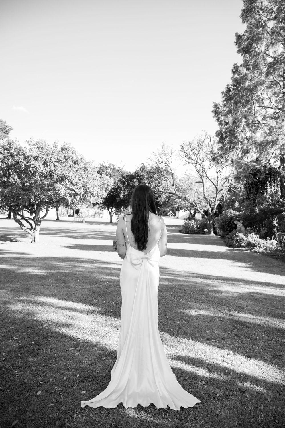 Black and white photo of bride in flowing dress standing alone in garden setting.