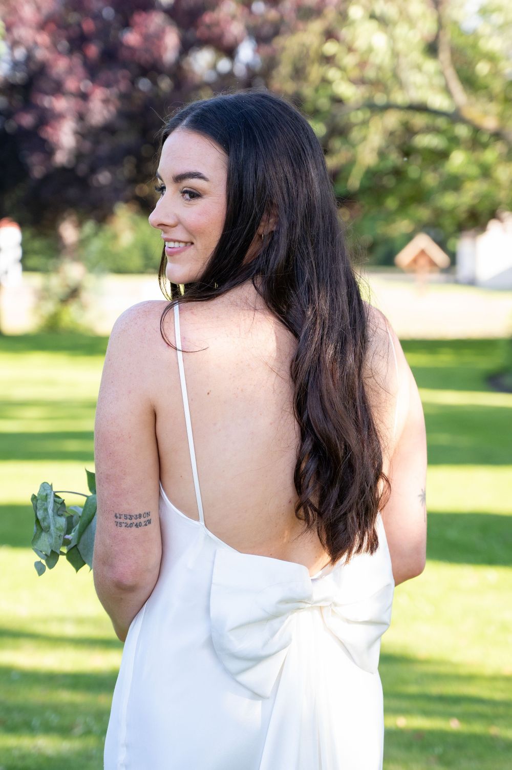 A woman in a white dress smiles while holding flowers in a sunny garden setting.