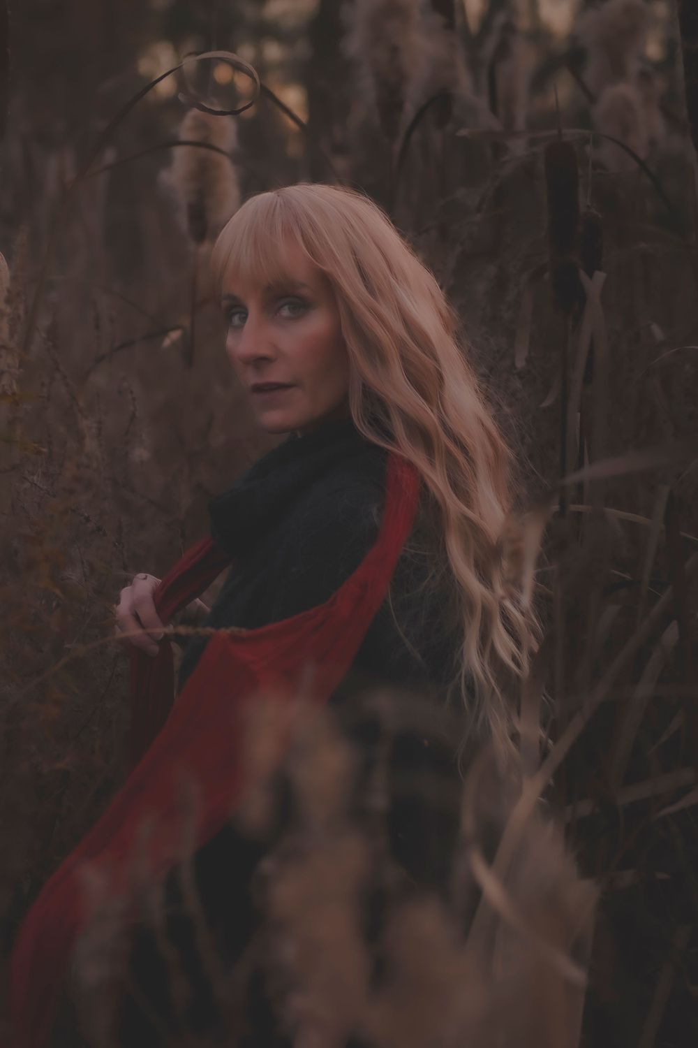 A person with long blonde hair wearing black and red clothing poses mysteriously in an autumn forest setting.