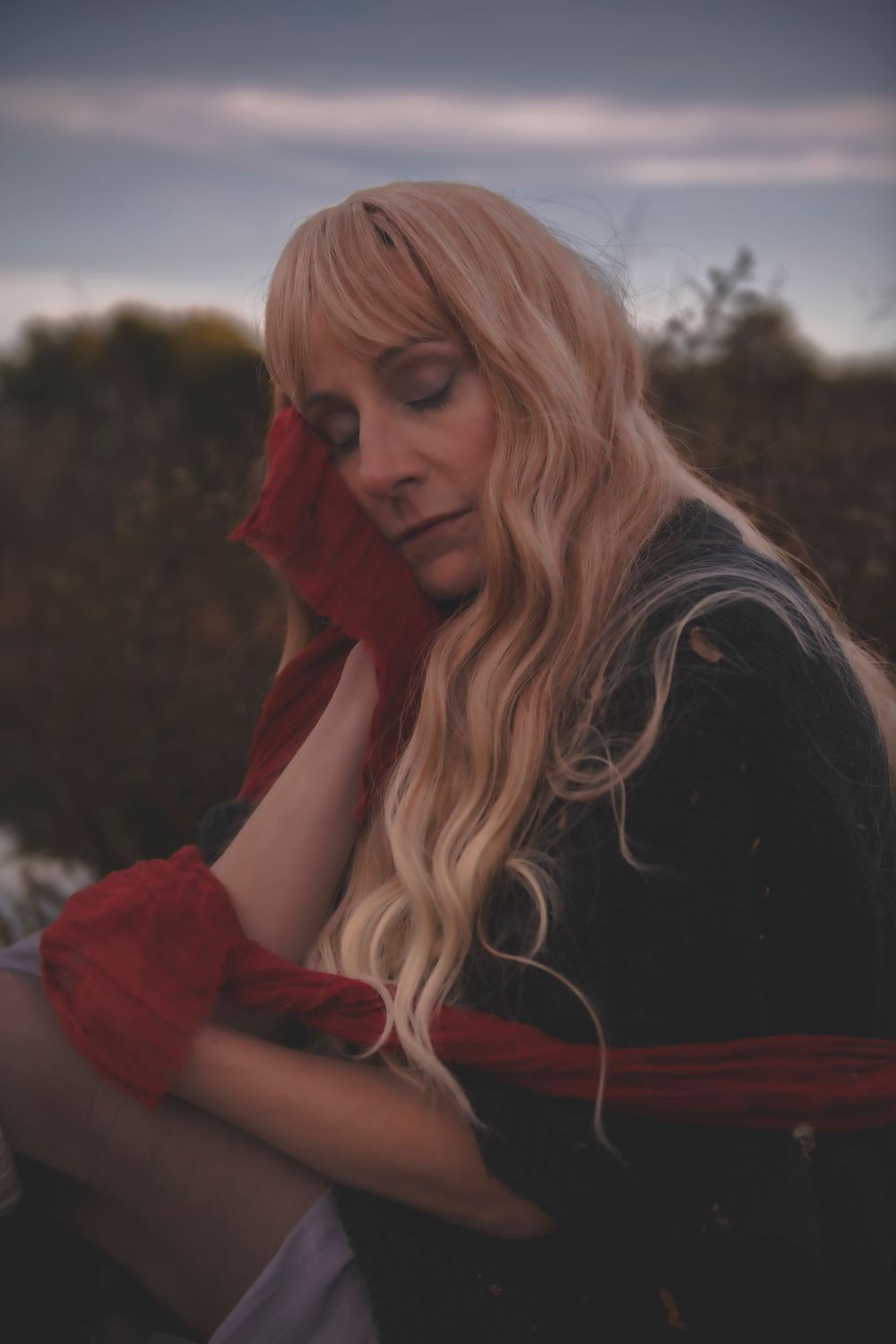Ethereal portrait features long blonde hair and red accents against dusky natural background.