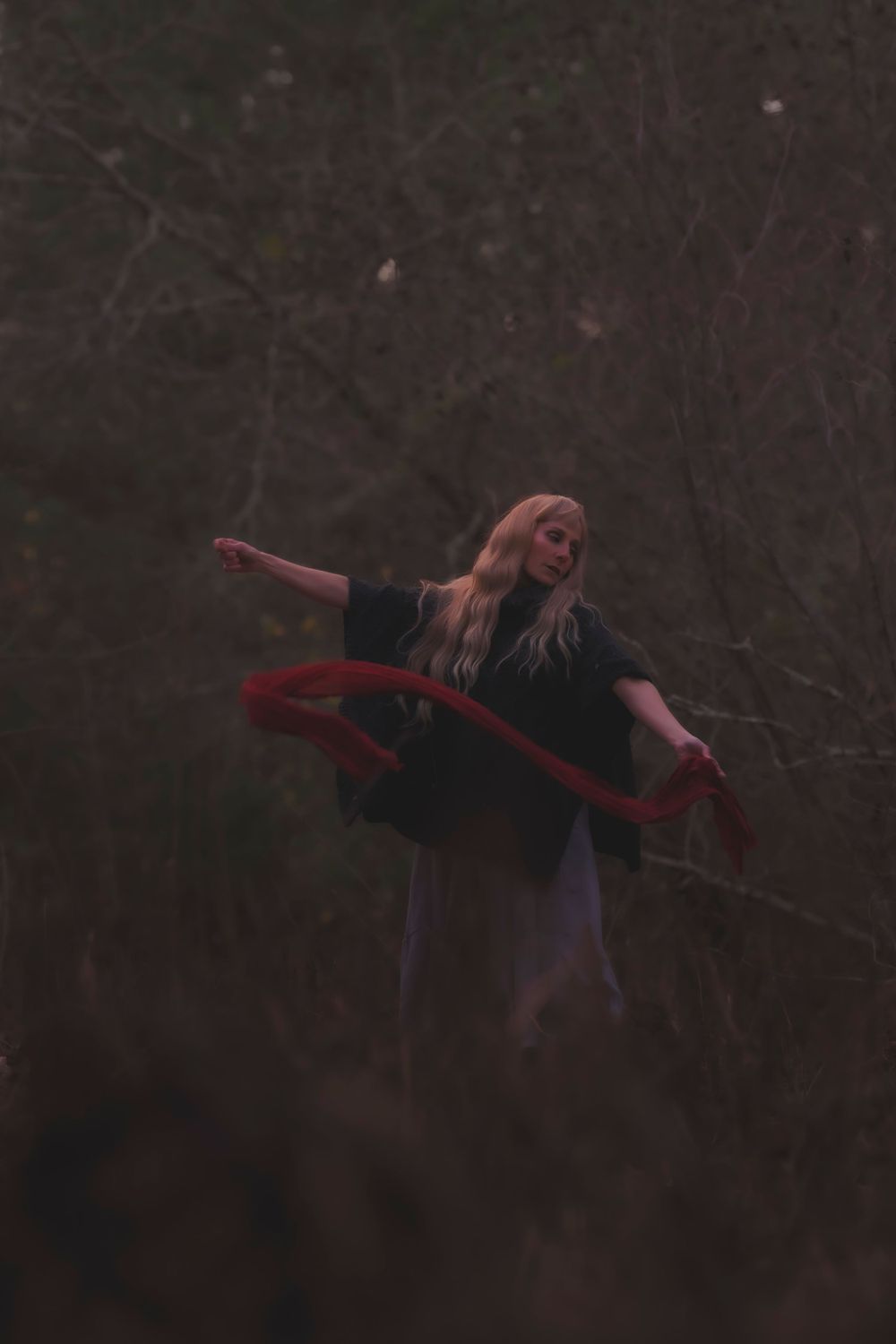 A figure in dark clothing dances with a flowing red scarf against a moody forest backdrop in dim lighting.