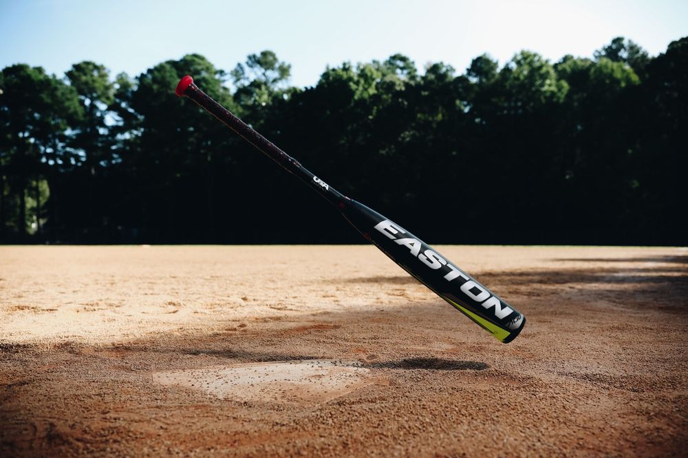 Easton baseball bat tossed in the air above a sandy baseball field with trees in background.