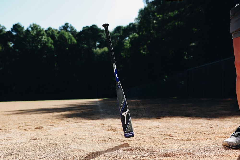 Black baseball bat standing upright in sandy infield dirt on a baseball field with trees behind.