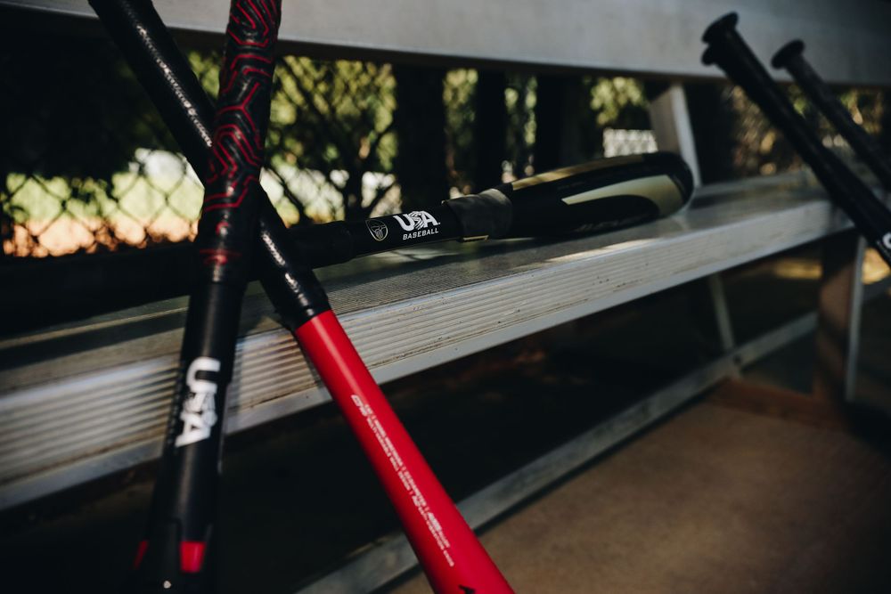 Red and black USA Baseball certified bats leaning against a dugout bench in low light.