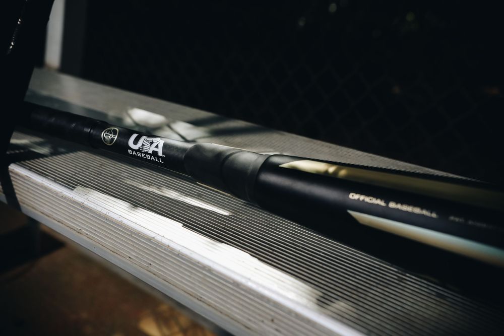 Close-up of a black USA Baseball certified official baseball bat resting on a dugout bench.