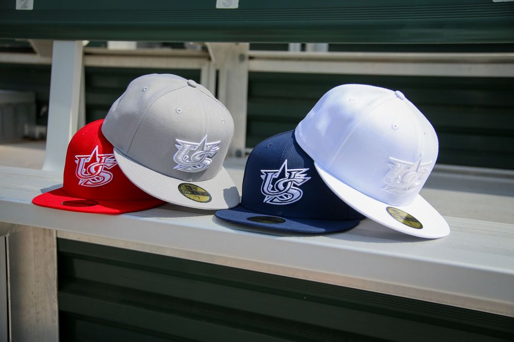 Four USA Baseball fitted caps in red, gray, navy, and white displayed on dugout bench.