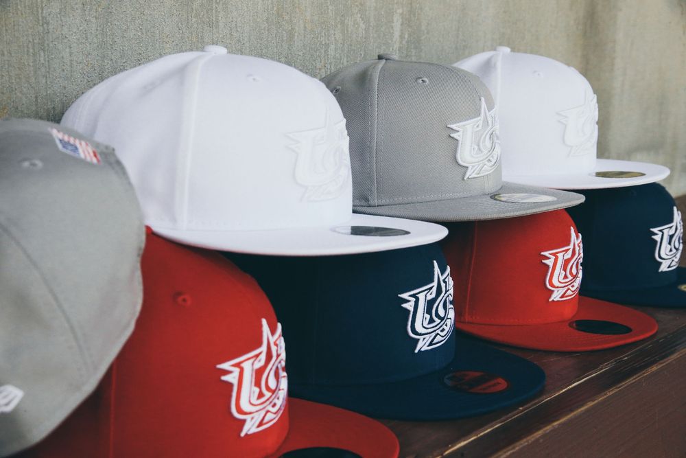 Row of USA Baseball New Era fitted caps in white, gray, navy, and red on bench.