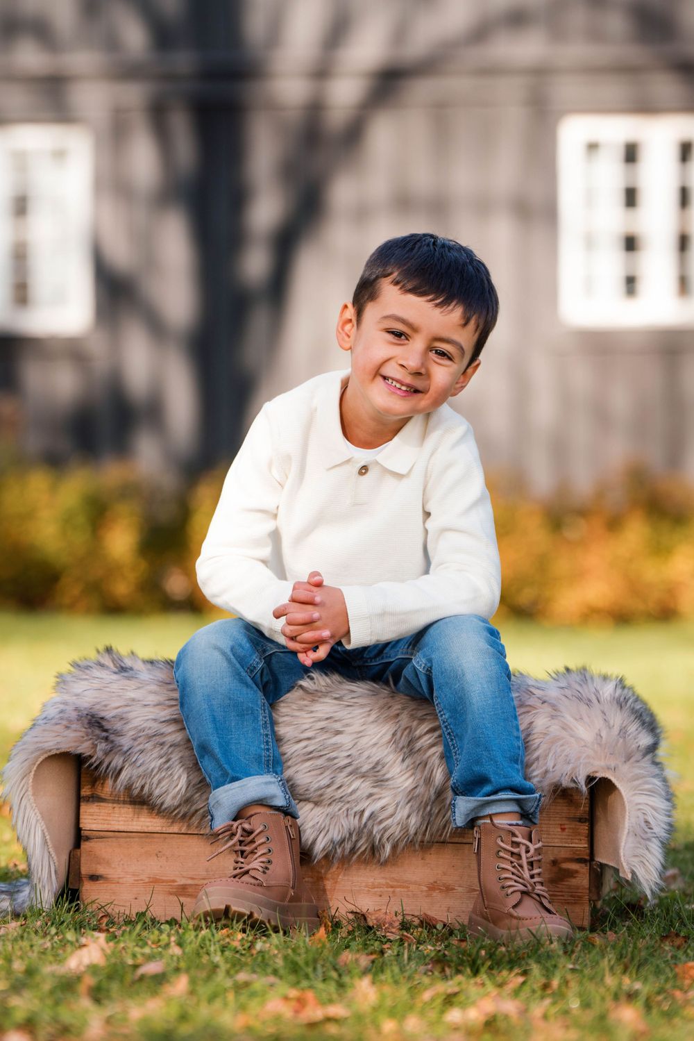 A child sits on a wooden crate with gray fur throw while smiling outdoors on grass with buildings in background.