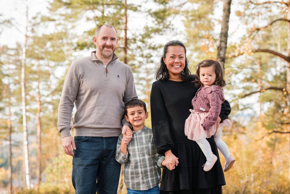 A happy family of four poses together outdoors among fall foliage and pine trees on a sunny autumn day.