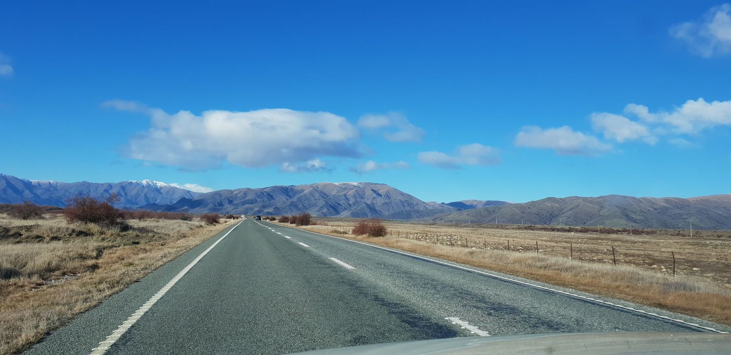 A long empty desert highway stretches towards distant mountains under a bright blue sky with scattered clouds.