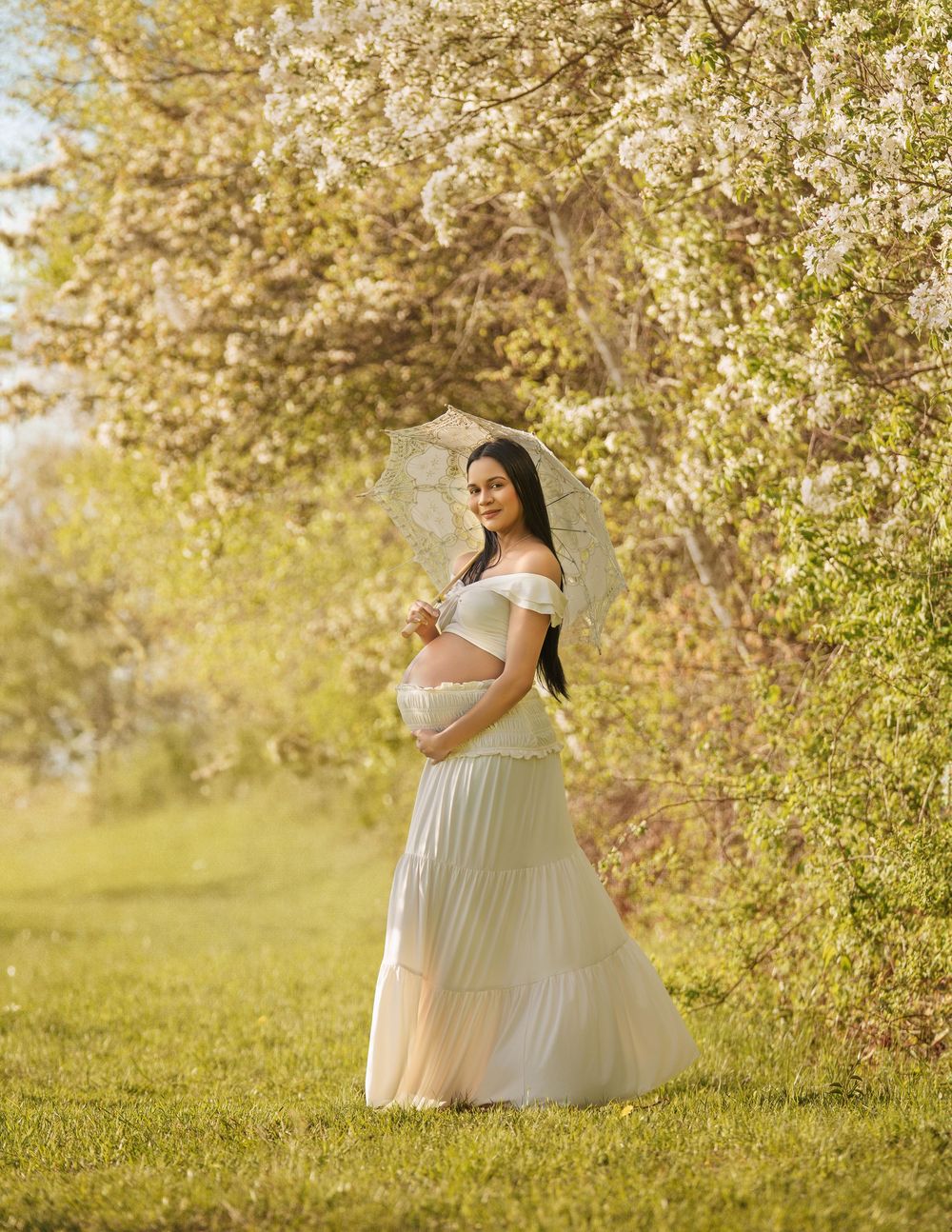 Outdoor maternity portrait in white dress among spring blossoms.