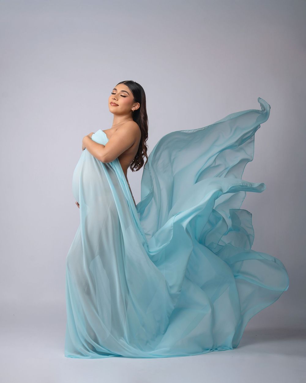 Woman in flowing light blue chiffon gown poses gracefully with fabric billowing dramatically against a soft gray studio background.