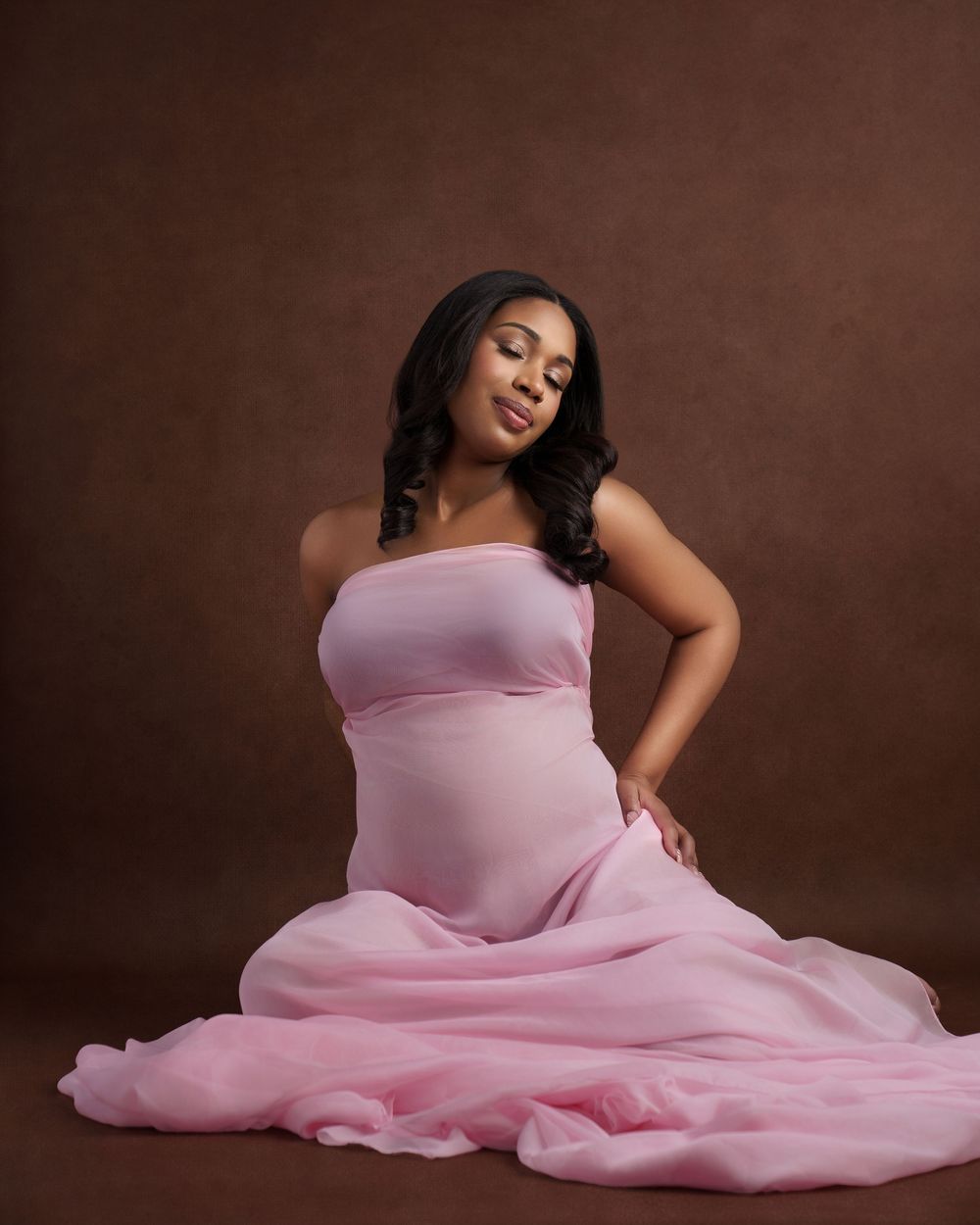 Elegant maternity photo featuring flowing pink gown against brown backdrop.